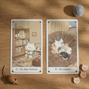 May include: Two rectangular tarot cards featuring cat illustrations. The left card, labeled II. The High Priestess, shows a white cat reading in a library. The right card, labeled III. The Empress, depicts a large cat with kittens on a sofa.