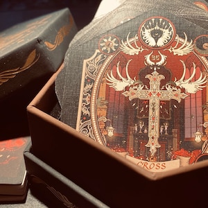 May include: A black box with a gold design on the lid is open, revealing a stack of cards with intricate designs. The top card features a gold cross with wings and the word "CROSS" printed below it.