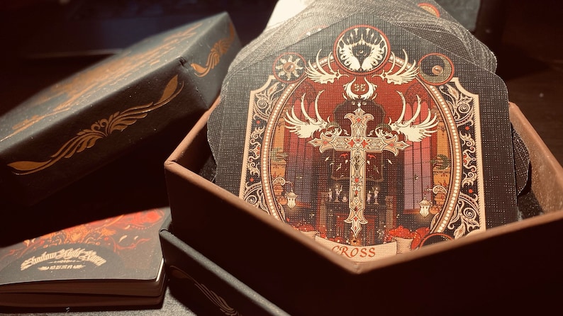 May include: A black box with a gold design on the lid is open, revealing a stack of cards with intricate designs. The top card features a gold cross with wings and the word "CROSS" printed below it.