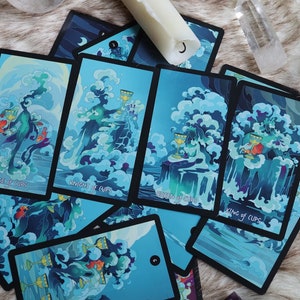 K&ouml;nnte beinhalten: Ein Satz Tarotkarten mit einem blauen und gr&uuml;nen Wasserthema. Die Karten liegen auf einer wei&szlig;en, flauschigen Oberfl&auml;che ausgebreitet. Die Karten zeigen den Page, den Ritter, die K&ouml;nigin und den K&ouml;nig der Kelche.