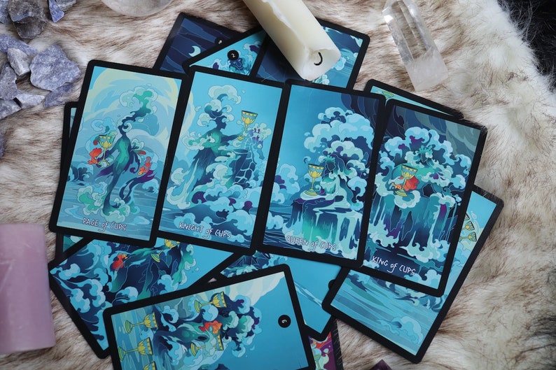 K&ouml;nnte beinhalten: Ein Satz Tarotkarten mit einem blauen und gr&uuml;nen Wasserthema. Die Karten liegen auf einer wei&szlig;en, flauschigen Oberfl&auml;che ausgebreitet. Die Karten zeigen den Page, den Ritter, die K&ouml;nigin und den K&ouml;nig der Kelche.