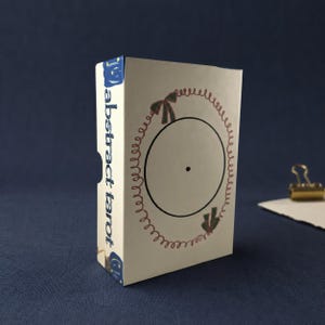 Puede incluir: Una caja rectangular de color blanco hueso para una baraja de cartas con la etiqueta "abstract tarot" en texto azul en el lateral. El frente presenta un círculo negro central con un pequeño punto, rodeado por un borde en espiral marrón decorado con dos lazos verdes.