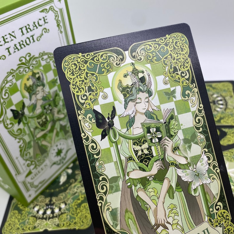 Może przedstawiać: Karta tarota z talii "Green Trace Tarot", z zieloną ilustracją ukoronowanej postaci trzymającej książkę i laskę z motylem. Karta ma czarną obw&oacute;dkę z zielonymi wzorami i tekstem "GREEN TRACE TAROT".