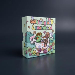 May include: A rectangular deck box for Whimsyland Lenormand cards featuring a light green background with a colourful illustration of two cartoon characters sitting in a polka-dot teacup surrounded by flowers, balloons, and confetti.