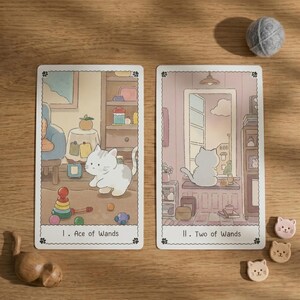 May include: Two rectangular tarot cards featuring cat illustrations. The left card, labeled I. Ace of Wands, shows a white cat with toys. The right card, labeled II. Two of Wands, shows a white cat looking out a window. Three small cat-shaped wooden tokens are nearby.