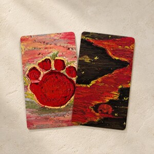 May include: Two rectangular, textured art pieces. One features a large red paw print on a pink and yellow background. The other has a dark, fiery landscape with red and yellow hues, and small white accents.