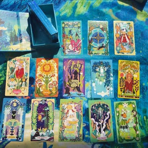 Puede incluir: Una baraja de cartas de tarot con ilustraciones coloridas, incluyendo im&aacute;genes de flores, &aacute;rboles y figuras. Las cartas est&aacute;n dispuestas sobre una superficie azul estampada, con una caja a juego en el fondo. La baraja es nueva.
