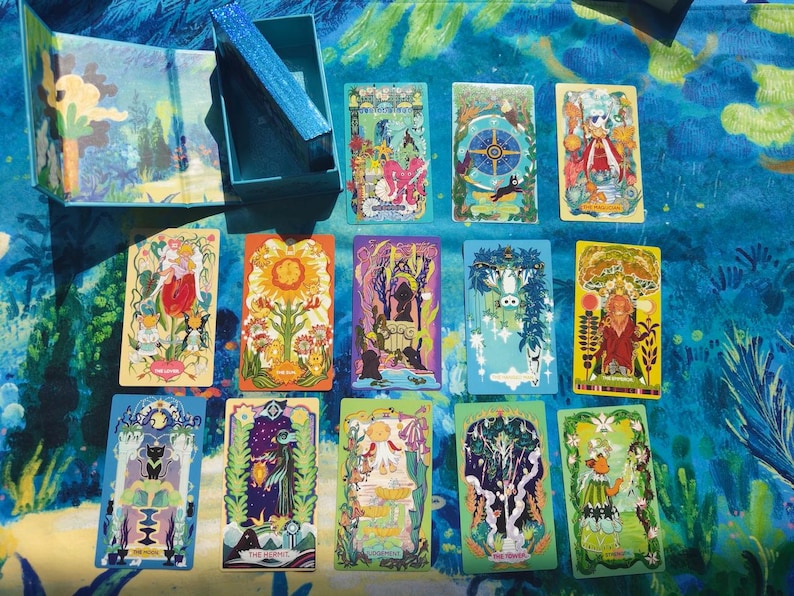 Puede incluir: Una baraja de cartas de tarot con ilustraciones coloridas, incluyendo im&aacute;genes de flores, &aacute;rboles y figuras. Las cartas est&aacute;n dispuestas sobre una superficie azul estampada, con una caja a juego en el fondo. La baraja es nueva.