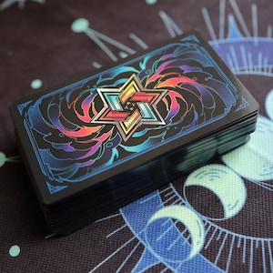 K&ouml;nnte beinhalten: Ein Kartenspiel mit schwarzem Hintergrund und einem farbenfrohen, geometrischen Design in der Mitte. Das Design zeigt eine Sternform mit mehreren Schichten und einem Regenbogen-Farbschema.