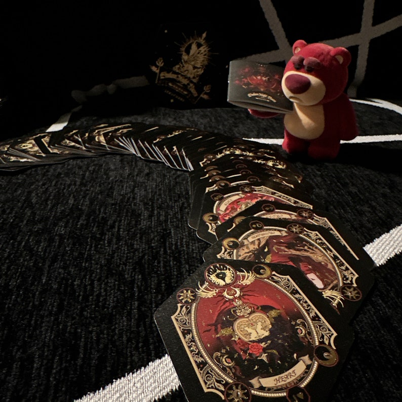 May include: A spread of tarot cards with a red and gold design. The cards are fanned out in a semi-circle on a black surface. A plush red teddy bear is holding a card in its paws.