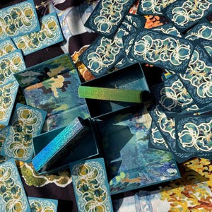 Puede incluir: Vista a&eacute;rea de un conjunto de cajas y tarjetas de color turquesa. Las tarjetas presentan un dise&ntilde;o floral blanco y dorado. Las cajas tienen un dise&ntilde;o de paisaje y acentos iridiscentes. Los art&iacute;culos est&aacute;n dispuestos sobre una superficie estampada.