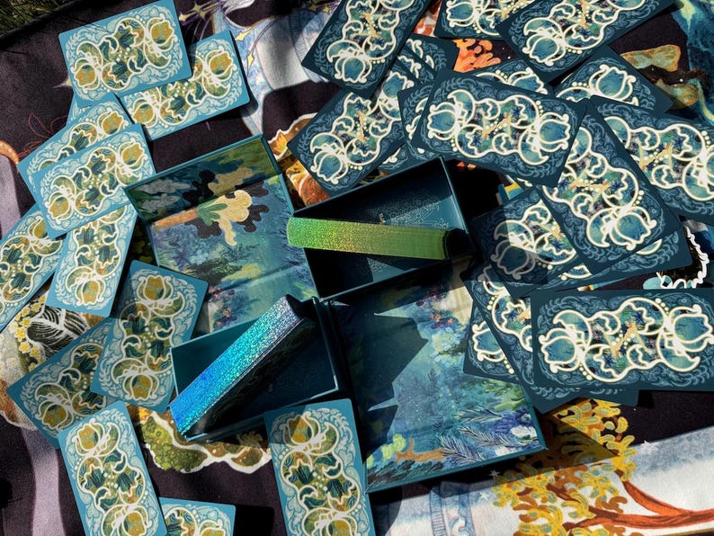 Puede incluir: Vista a&eacute;rea de un conjunto de cajas y tarjetas de color turquesa. Las tarjetas presentan un dise&ntilde;o floral blanco y dorado. Las cajas tienen un dise&ntilde;o de paisaje y acentos iridiscentes. Los art&iacute;culos est&aacute;n dispuestos sobre una superficie estampada.