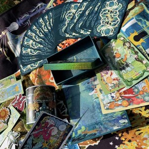 Puede incluir: Una colecci&oacute;n de cartas del tarot, una caja y una lata decorativa. Las cartas son de color verde azulado con dise&ntilde;os florales blancos. La caja es de color verde azulado con un interior hologr&aacute;fico. La lata presenta un dise&ntilde;o de hongo. Varias cartas est&aacute;n dispersas.