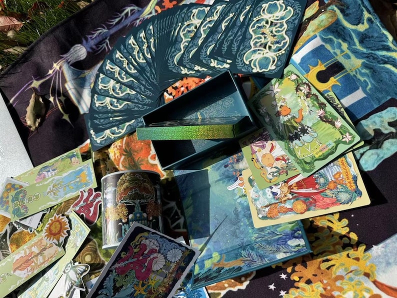 Puede incluir: Una colecci&oacute;n de cartas del tarot, una caja y una lata decorativa. Las cartas son de color verde azulado con dise&ntilde;os florales blancos. La caja es de color verde azulado con un interior hologr&aacute;fico. La lata presenta un dise&ntilde;o de hongo. Varias cartas est&aacute;n dispersas.