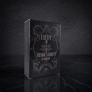 Op de afbeelding: Een zwart rechthoekig tarotdeckdoosje met de witte tekst Elegy of Black Lichen Tarot. Het ontwerp op de voorzijde toont een gestileerde bloem in het midden, omringd door gedetailleerde illustraties van doornige ranken en kleine vierpuntige sterren.