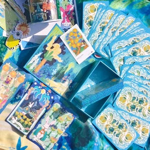 Puede incluir: Un conjunto de cartas de tarot con ilustraciones coloridas, presentado con una caja a juego. Las cartas y la caja tienen una combinaci&oacute;n de colores azul y blanco, con ilustraciones detalladas. Las cartas est&aacute;n dispuestas sobre una superficie estampada en azul y blanco.