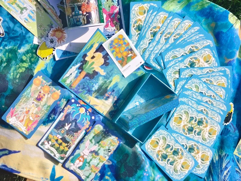 Puede incluir: Un conjunto de cartas de tarot con ilustraciones coloridas, presentado con una caja a juego. Las cartas y la caja tienen una combinaci&oacute;n de colores azul y blanco, con ilustraciones detalladas. Las cartas est&aacute;n dispuestas sobre una superficie estampada en azul y blanco.