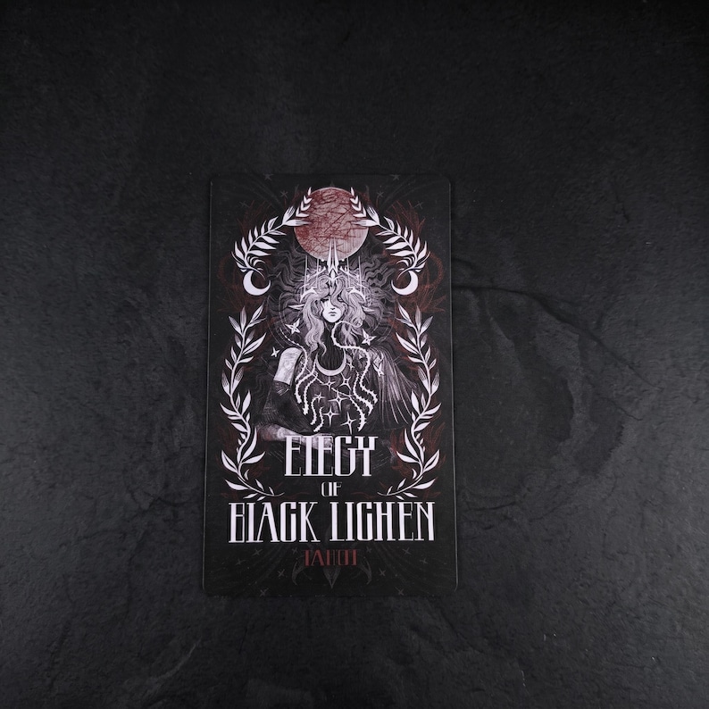 Op de afbeelding: Een rechthoekige kaart met een donkere illustratie van een figuur met lang haar, een kroon en sterachtige patronen. De tekst luidt ELEGY OF BLACK LIGHEN TAHITI. Het ontwerp is omlijst door witte bladeren en een rode maan op een zwarte achtergrond.