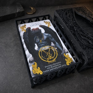 Puede incluir: Una baraja de cartas de tarot en un soporte de metal negro. La carta superior muestra una figura con alas oscuras, un collar de cruz dorada y un sello circular dorado. Texto: The Devil LUCIFER, PRIDE, Provides liberation and illumination. Incites jealousy and envy.