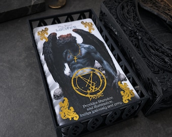 May include: A deck of tarot cards in a black metal holder. The top card features a dark-winged figure, a gold cross necklace, and a gold circular sigil. Text reads: The Devil LUCIFER, PRIDE, Provides liberation and illumination. Incites jealousy and envy.