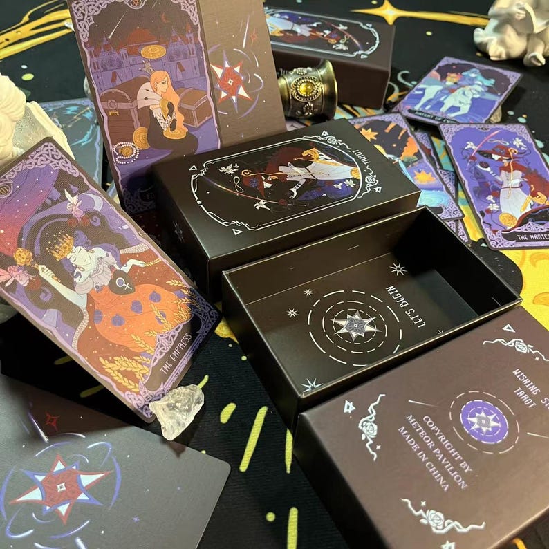 Op de afbeelding: Een zwarte tarotdeckdoos met een gouden sterontwerp en de tekst "Let's Begin". De doos is open en laat de kaarten zien die er in zitten, met kleurrijke illustraties en tekst zoals "The Empress" en "The Magic".