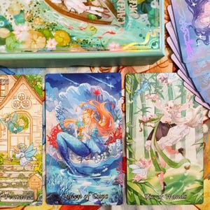 May include: Three tarot cards with illustrations of fairies. The card on the left is the Ten of Pentacles, the middle card is the Queen of Cups, and the card on the right is the Six of Wands.