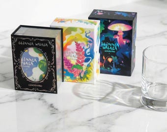 May include: Three illustrated boxes with the text "Glimmer Walker" and "Lenormand". The boxes feature vibrant designs of plants, mushrooms, and abstract shapes. A clear glass is positioned to the right of the boxes.