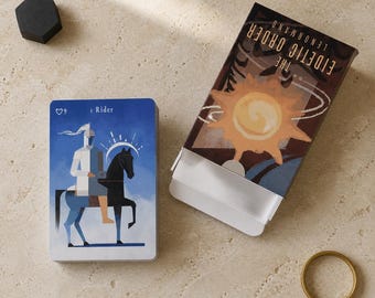 May include: A deck of The Eidetic Order Lenormand cards featuring a Rider card with a knight on a black horse against a blue sky. The box displays a sun illustration and the text The Eidetic Order Lenormand. A black hexagon and gold ring are present.