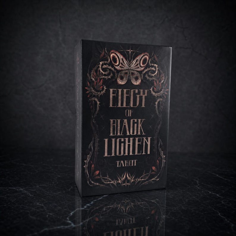 Op de afbeelding: Een rechthoekige tarotkaartendoos in zwart met koperkleurige letters die Elegy of Black Lichen Tarot spellen. De voorkant bevat een gedetailleerde illustratie van een vlinder omringd door doornige wijnstokken en gebladerte in kopertinten.