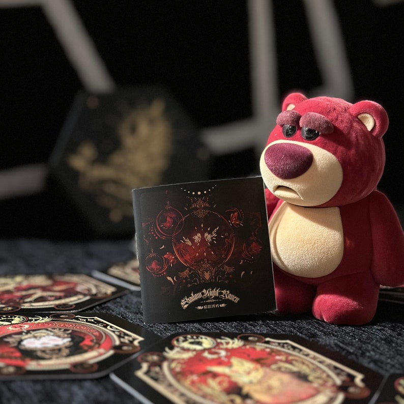 May include: A red plush toy bear with a sad expression is holding a small black booklet with a red and gold design. The booklet has the text "Shadow Fight Raven" on the front.