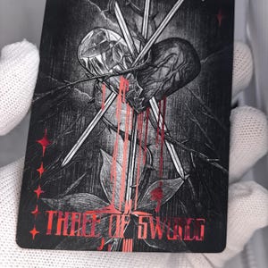 Op de afbeelding: Een rechthoekige tarotkaart met een zwart-witte illustratie van een hart doorboord door drie zwaarden, omringd door doornige wijnstokken. Het ontwerp bevat rode metalen accenten, bloedachtige druppels en de tekst Three of Swords in het rood.