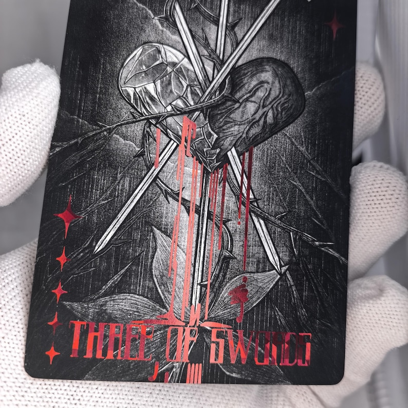 Op de afbeelding: Een rechthoekige tarotkaart met een zwart-witte illustratie van een hart doorboord door drie zwaarden, omringd door doornige wijnstokken. Het ontwerp bevat rode metalen accenten, bloedachtige druppels en de tekst Three of Swords in het rood.