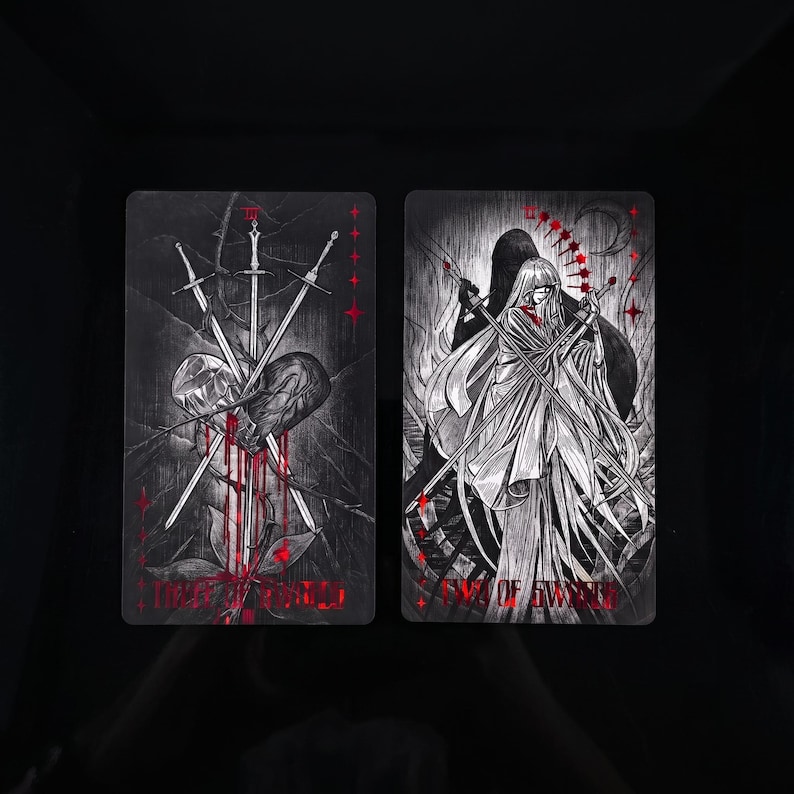 Op de afbeelding: Twee rechthoekige tarotkaarten met zwart-witte illustraties en rode accenten. De linkerkaart toont drie zwaarden die een hart doorboren met de tekst Three of Swords. De rechterkaart toont een figuur met de tekst Two of Swords.