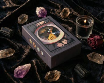 Op de afbeelding: Een tarotkaartdeksel met een oogontwerp, de tekst "THE SOUL'S HAVEN" en kleurrijke illustraties. De doos is omgeven door kristallen, een brandende kaars en een gedroogde roos op een donkere, getextureerde ondergrond.