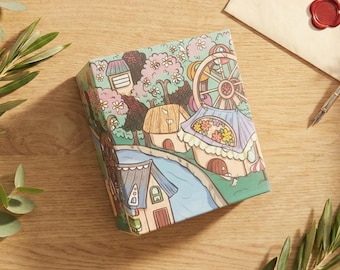 May include: A square box featuring a colorful illustration of a whimsical village with small houses, a Ferris wheel, and a winding blue river. The artwork includes pastel-toned trees, flowers, and detailed architectural elements on a light green background.