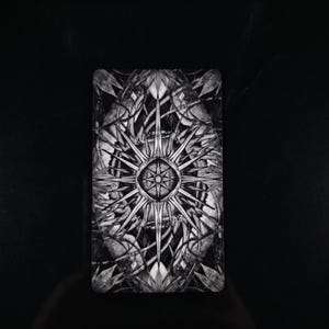 Op de afbeelding: Een rechthoekige tarotkaart met een zwart-wit symmetrisch ontwerp. Het midden toont een stervormig patroon met vele punten, omringd door complexe, vloeiende botanische motieven en bladvormige figuren die naar de randen toe uitlopen.