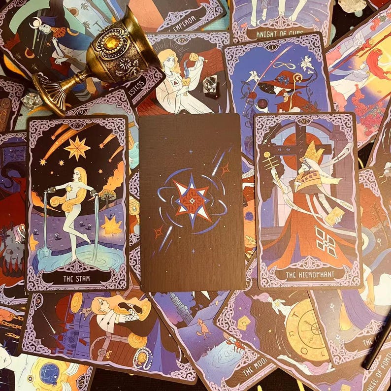 Op de afbeelding: Een verzameling tarotkaarten met kleurrijke illustraties en gouden accenten. De kaarten bevatten afbeeldingen van de Ster, de Hi&euml;rofant, de Keizer, de Ridder van Bekers en de Maan. De kaarten liggen op een donker oppervlak.