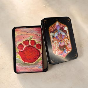 May include: Two black rectangular tins, one open, displaying artwork. The open tin reveals a vibrant red paw print on a textured background. The closed tin features a colorful illustration of a deity. The tins are on a neutral surface.