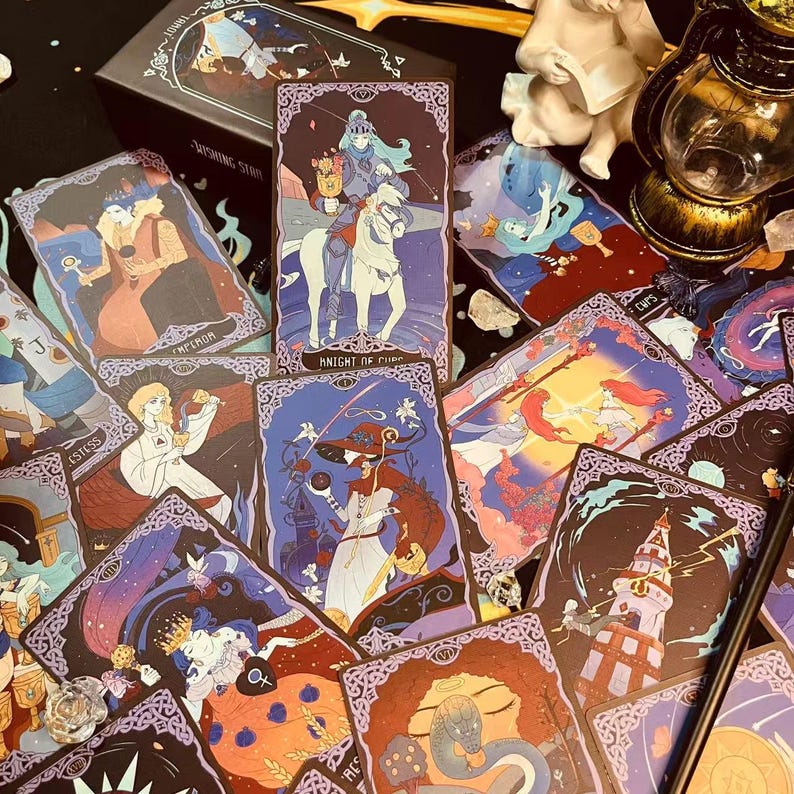 Op de afbeelding: Een set tarotkaarten met illustraties van mensen en dieren in een fantasiewereld. De kaarten hebben een donkere achtergrond met paarse en gouden accenten. De kaarten liggen op een tafel met een lamp en andere objecten op de achtergrond.