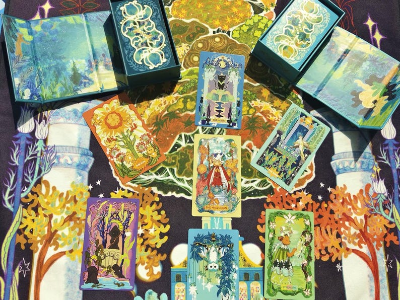 Puede incluir: Un conjunto de cartas de tarot con ilustraciones coloridas, que incluyen im&aacute;genes de un girasol, una figura con una t&uacute;nica roja y varias escenas. Las cartas est&aacute;n dispuestas sobre una tela estampada, con dos cajas y un folleto plegado.