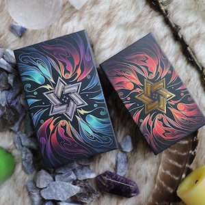 K&ouml;nnte beinhalten: Zwei schwarze Boxen mit bunten, wirbelnden Designs und einem goldenen Stern in der Mitte. Die Boxen befinden sich auf einer braunen, flauschigen Oberfl&auml;che mit kleinen grauen Steinen und einer Feder.