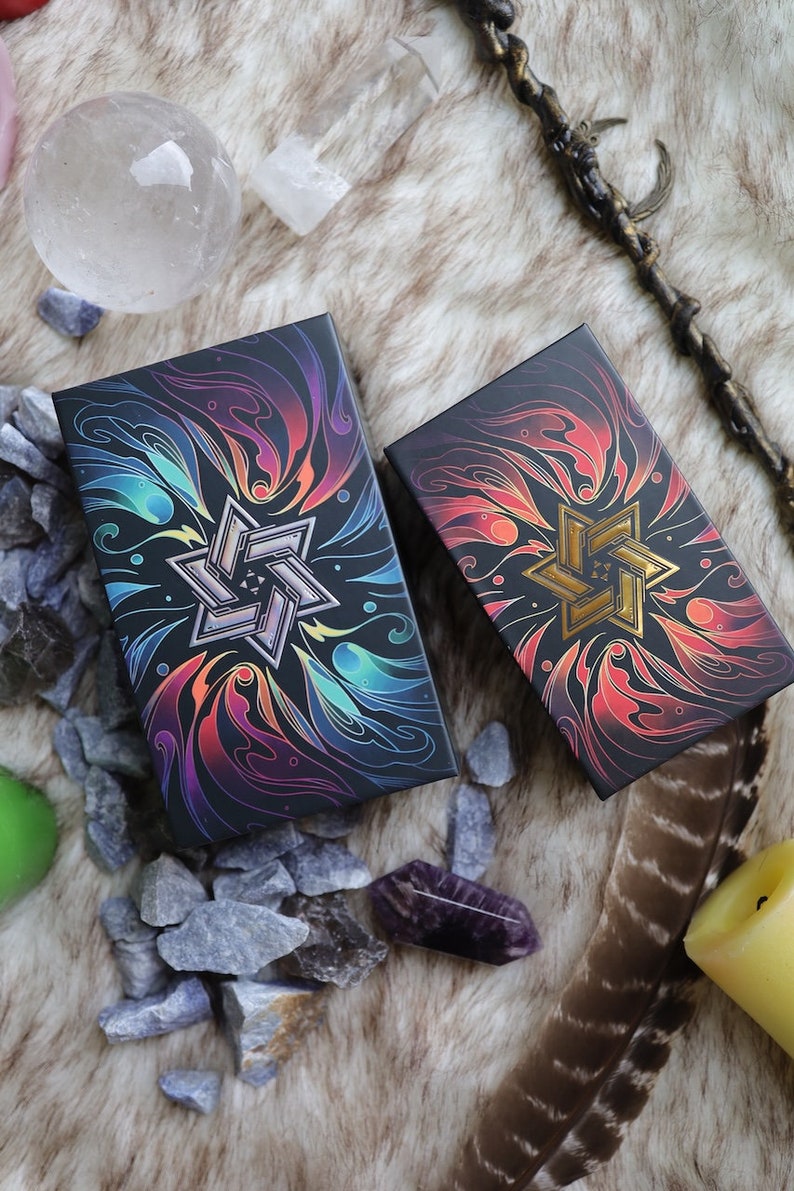 K&ouml;nnte beinhalten: Zwei schwarze Boxen mit bunten, wirbelnden Designs und einem goldenen Stern in der Mitte. Die Boxen befinden sich auf einer braunen, flauschigen Oberfl&auml;che mit kleinen grauen Steinen und einer Feder.