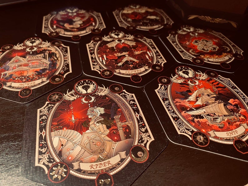 May include: A set of tarot cards with intricate gold and black designs. The cards feature illustrations of a house, clouds, a ship, and a rider on horseback. The cards are set against a dark red background.