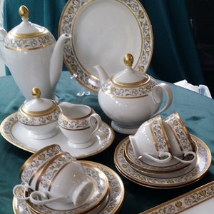 Puede incluir: Un juego de té de porcelana blanca con ribetes dorados y detalles florales. El juego incluye una tetera, una lechera, un azucarero, tazas, platillos y una fuente. Los ribetes dorados y el diseño floral añaden un toque elegante.