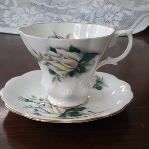May include: A white teacup and saucer with a gold rim and a floral design featuring a white rose with green leaves.