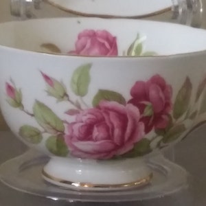 May include: A white teacup and saucer featuring a pink rose floral pattern. The teacup has a pedestal base and a gold trim. The saucer also has a gold trim. A traditional tea set, perfect for serving tea. The teacup and saucer are made of porcelain.