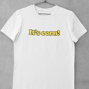 Può includere: T-shirt bianca con la scritta "It's corn!" in giallo, bordata di giallo più scuro. Il testo ha un aspetto strutturato, simile al mais. La maglietta è appesa a una gruccia nera su sfondo grigio.