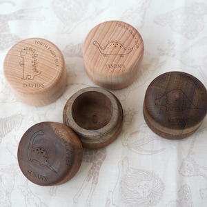 May include: Four round wooden tooth fairy boxes with lids. Three boxes are closed, showing engraved dinosaur illustrations and the text "TOOTH FAIRY BOX" along with names: David's, Jason's, and Kevin's. One dark wood box is open, showing its interior.
