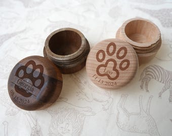 May include: Wooden keepsake boxes with paw print designs. One box is dark brown with the name "Georgia" and date "08.15.2024" engraved. Another is light wood with "Leo" and date "11.13.2024". The boxes are open and closed, showcasing the engraved paw print lids.
