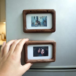 Könnte beinhalten: Zwei rechteckige magnetische Fotorahmen aus dunklem Holz, die an einem Kühlschrank befestigt sind. Der obere Rahmen trägt die Gravur "Our Special Day" und "06.08.2027". Der untere Rahmen trägt die Gravur "Olivia & Mia" unter einem Foto.